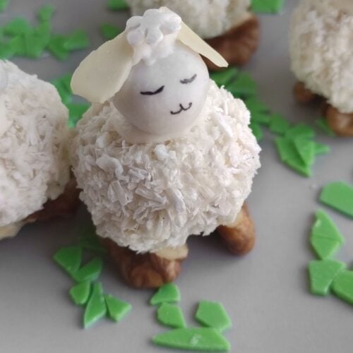 A dessert shaped like a sheep, featuring a coconut-covered body, almond head, and small eyes and mouth. It stands on walnuts with green candy pieces scattered around.