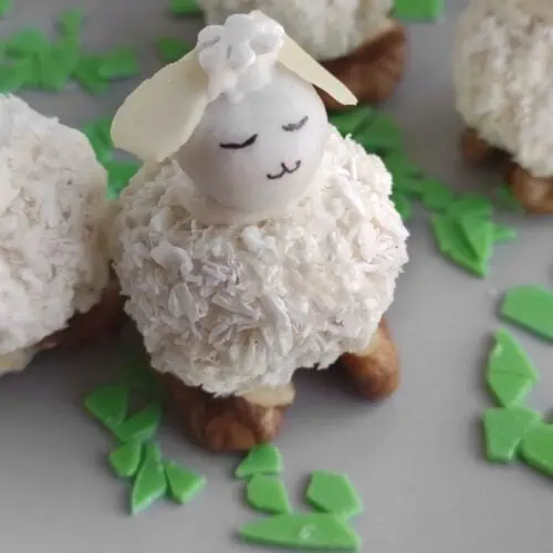 A dessert shaped like a sheep, featuring a coconut-covered body, almond head, and small eyes and mouth. It stands on walnuts with green candy pieces scattered around.
