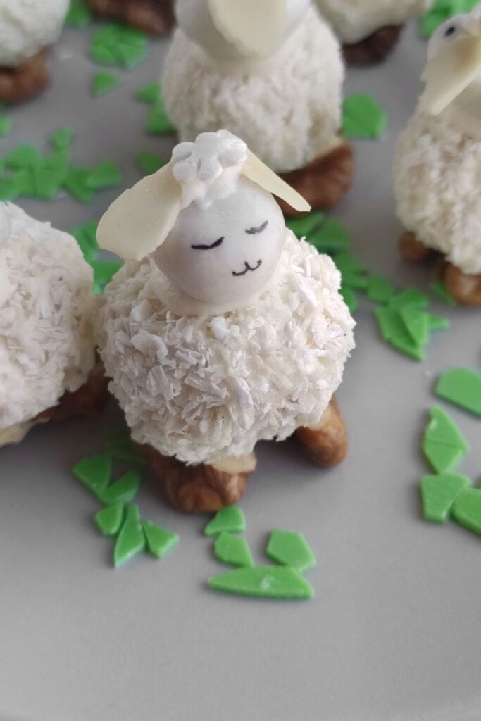 A dessert shaped like a sheep, featuring a coconut-covered body, almond head, and small eyes and mouth. It stands on walnuts with green candy pieces scattered around.