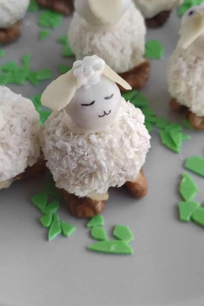 A dessert shaped like a sheep, featuring a coconut-covered body, almond head, and small eyes and mouth. It stands on walnuts with green candy pieces scattered around.