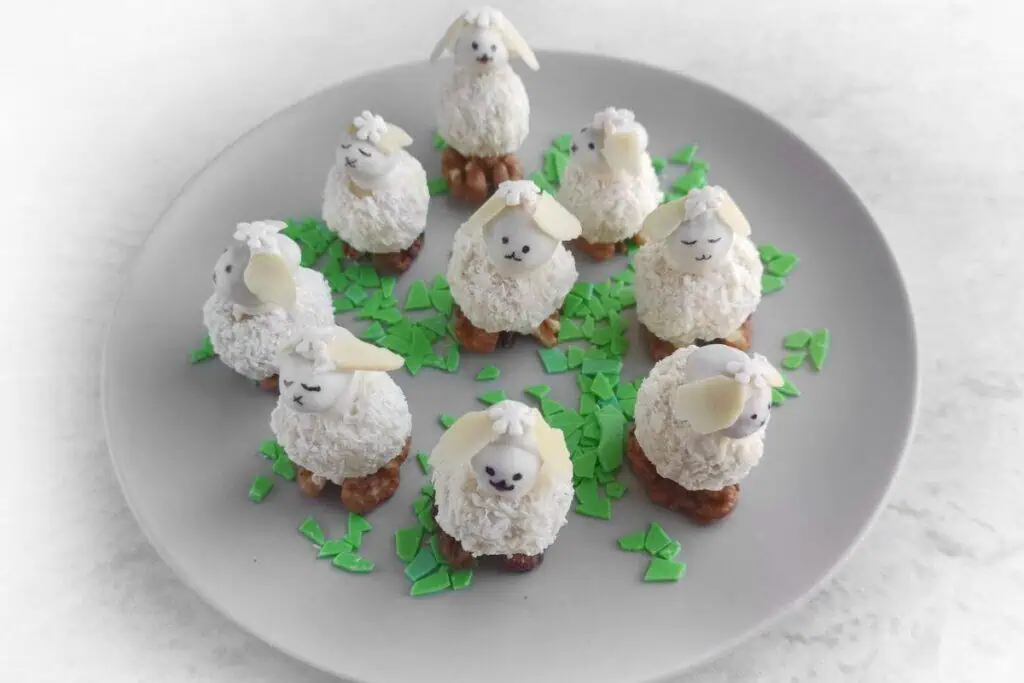 Nine sheep-shaped coconut desserts with almond ears and chocolate chip eyes on a plate with green sugar sprinkles.