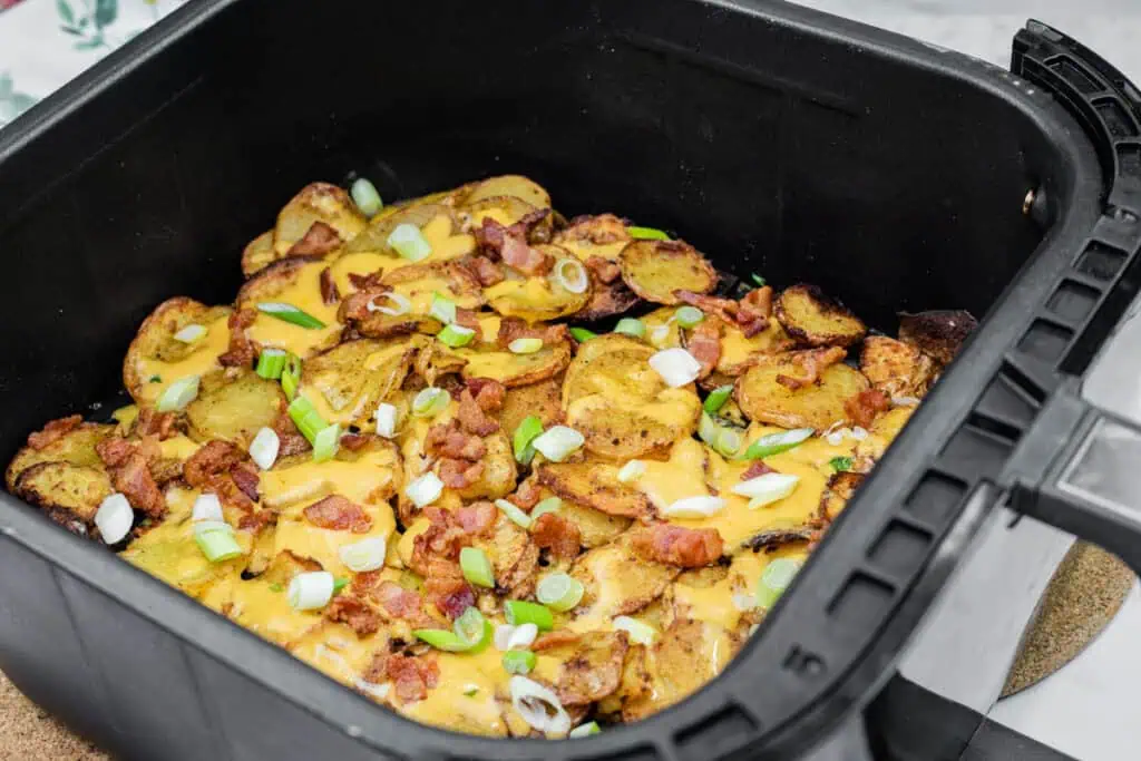 Cooked potato slices topped with melted cheese, bacon bits, and chopped green onions in a black square pan.