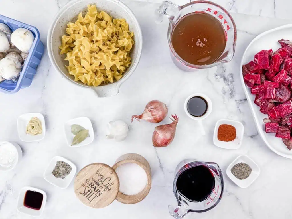 An array of ingredients for Beef Stroganoff Soup, including pasta, mushrooms, beef, broth, wine, shallots, garlic, spices, and seasonings, are displayed on a white marble surface.