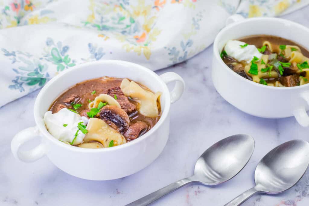 Two white bowls of Beef Stroganoff Soup with pasta, garnished with herbs and sour cream, are placed on a marble surface alongside two spoons and a floral-patterned cloth.