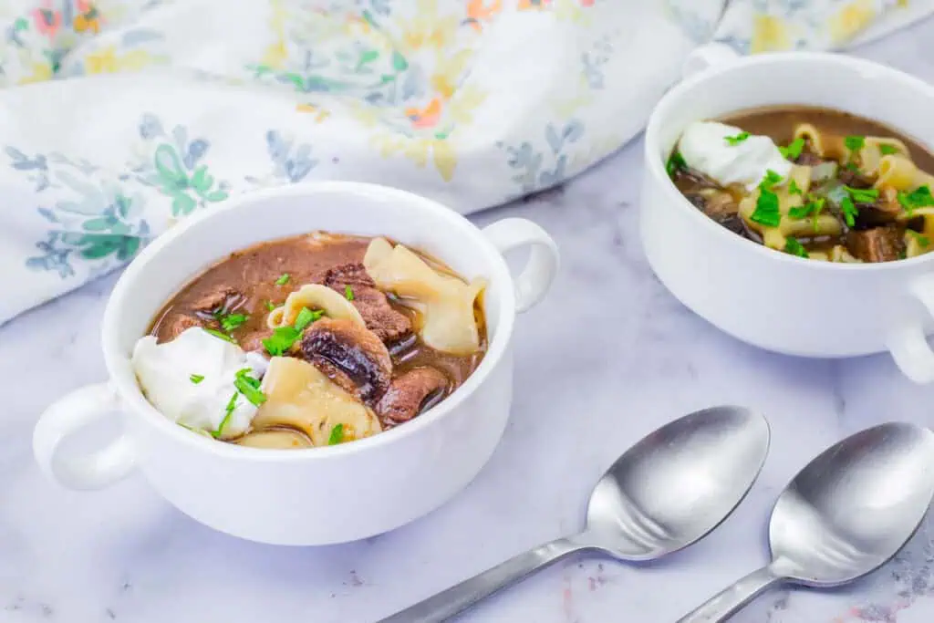 Two white bowls of Beef Stroganoff Soup with pasta, garnished with herbs and sour cream, are placed on a marble surface alongside two spoons and a floral-patterned cloth.