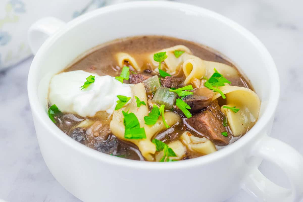 A white bowl of Beef Stroganoff Soup topped with sour cream and chopped parsley.