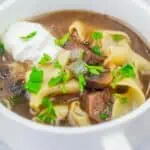 A bowl of beef stroganoff soup topped with a dollop of sour cream and garnished with parsley.