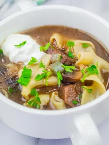 A bowl of beef stroganoff soup topped with a dollop of sour cream and garnished with parsley.