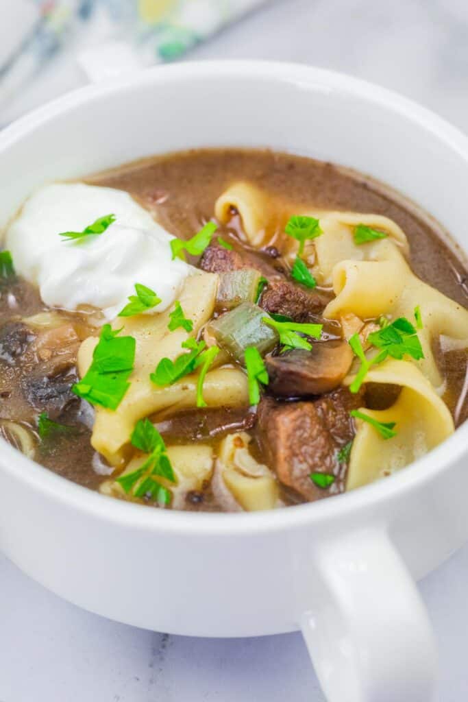 A bowl of beef stroganoff soup topped with a dollop of sour cream and garnished with parsley.