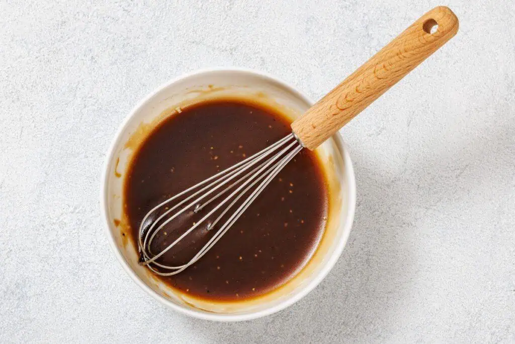 A white bowl with brown sauce and a whisk with a wooden handle resting inside, placed on a light surface.