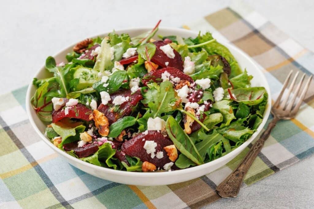 A colorful salad with mixed greens, sliced beets, walnuts, and crumbled feta in a white bowl on a plaid cloth, accompanied by a vintage fork.