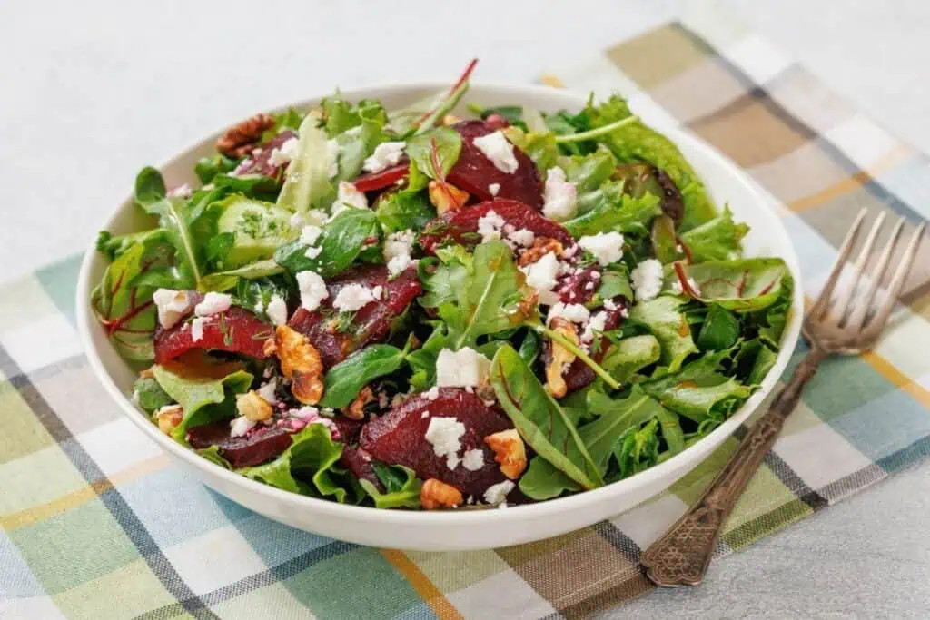 A colorful salad with mixed greens, sliced beets, walnuts, and crumbled feta in a white bowl on a plaid cloth, accompanied by a vintage fork.