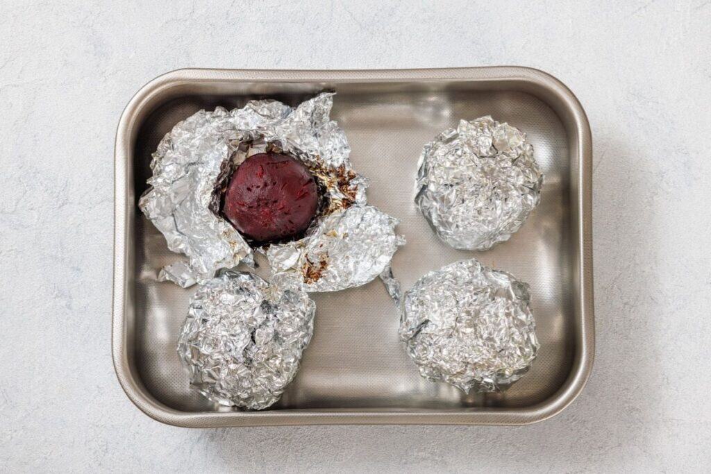 Four foil-wrapped items in a baking pan, with one partially unwrapped to reveal a cooked beet.