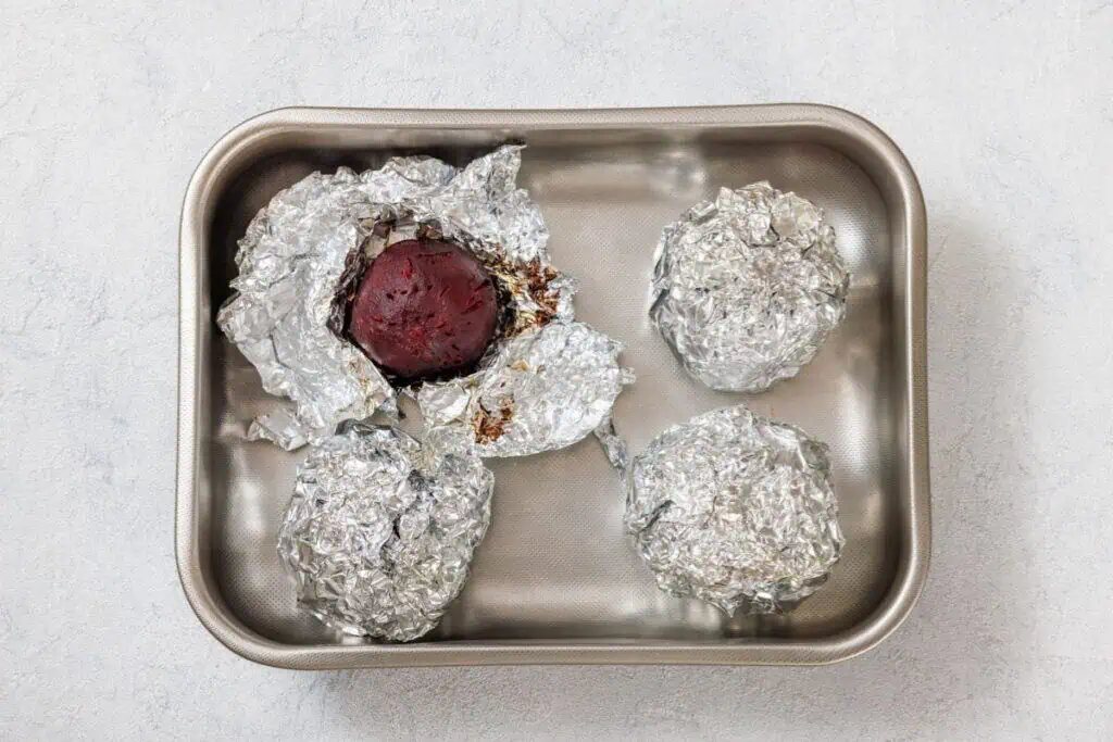 Four foil-wrapped items in a baking pan, with one partially unwrapped to reveal a cooked beet.