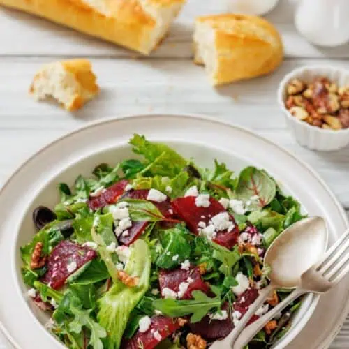 A salad with mixed greens, beets, feta, and walnuts on a plate. Beside it are a baguette, salt and pepper shakers, and a dish of jam.
