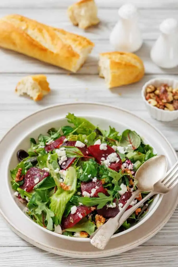 A salad with mixed greens, beets, feta, and walnuts on a plate. Beside it are a baguette, salt and pepper shakers, and a dish of jam.