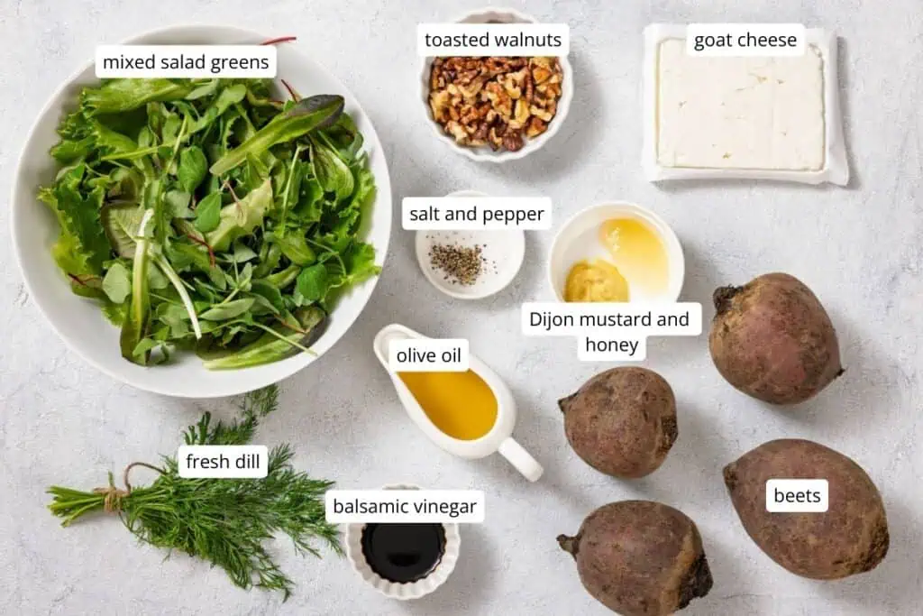 Ingredients on a countertop include mixed salad greens, fresh dill, balsamic vinegar, olive oil, toasted walnuts, salt and pepper, Dijon mustard and honey, goat cheese, and beets.