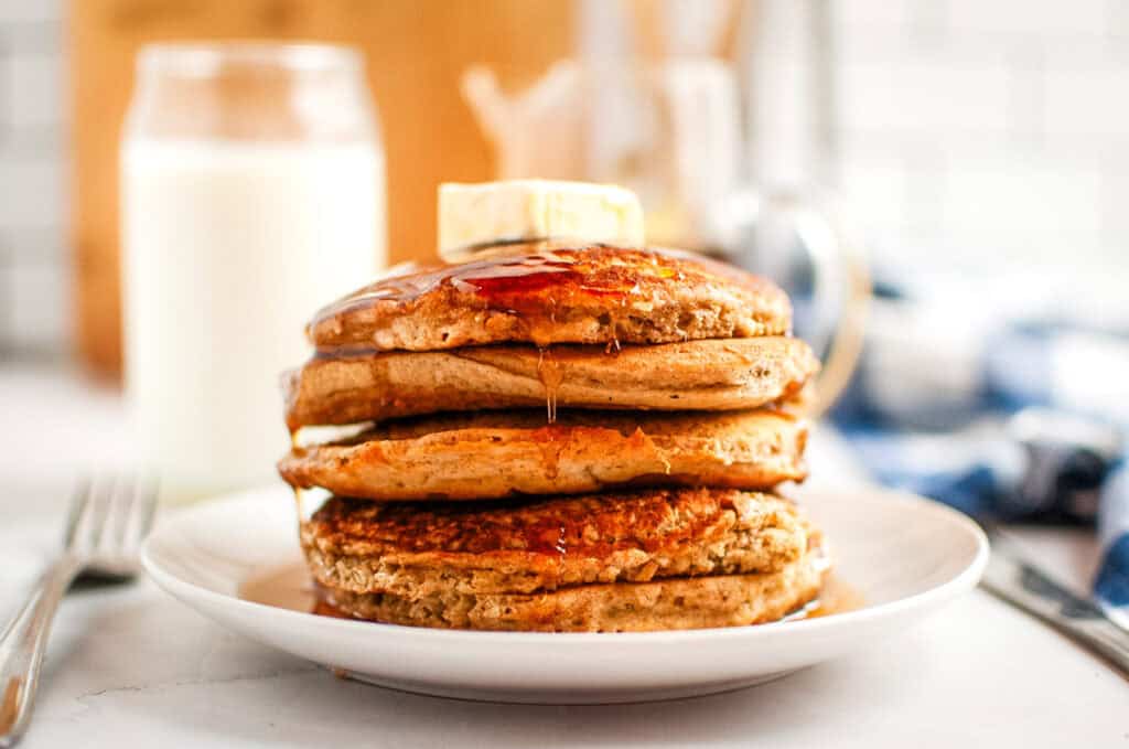 A stack of four Buttermilk Oatmeal Pancakes topped with a pat of butter and syrup on a white plate. A glass of milk is in the background along with a fork and a blurred blue item.