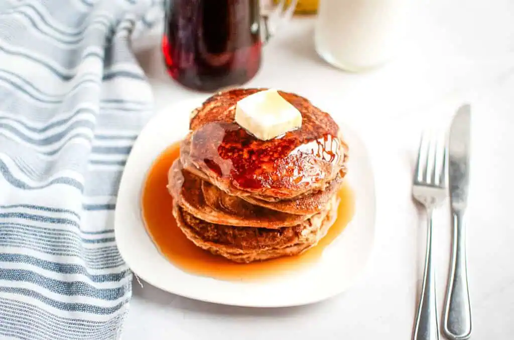 Stack of Buttermilk Oatmeal Pancakes with syrup and a pat of butter on top, served on a white plate, next to a blue-striped cloth and cutlery.