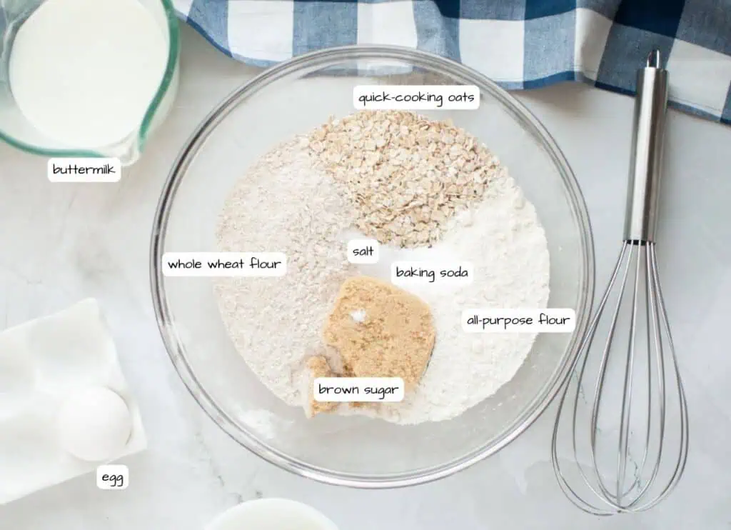 A bowl with labeled ingredients for Buttermilk Oatmeal Pancakes: quick-cooking oats, whole wheat flour, salt, baking soda, all-purpose flour, and brown sugar. Nearby are a whisk, egg, and buttermilk.