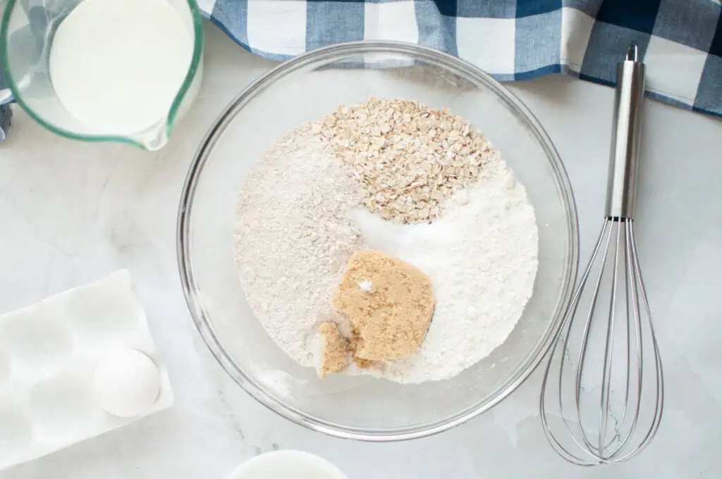 Bowl with flour, oats, brown sugar, and salt. Nearby are a whisk, milk in a pitcher, a blue checkered cloth, and an egg in an egg tray on a white surface.