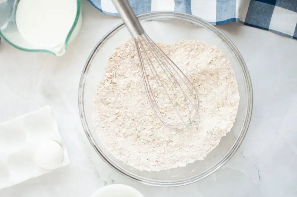 A whisk in a glass bowl containing flour on a marble countertop. Nearby are a measuring cup with milk, an egg, an egg tray, and a blue and white checkered cloth.