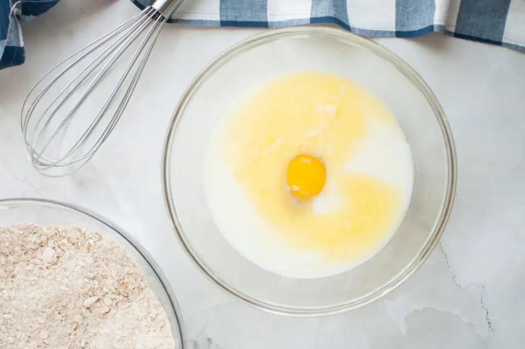 A bowl containing milk, melted butter, and an egg on a marble surface, next to a whisk and a bowl of dry ingredients with a blue and white cloth.