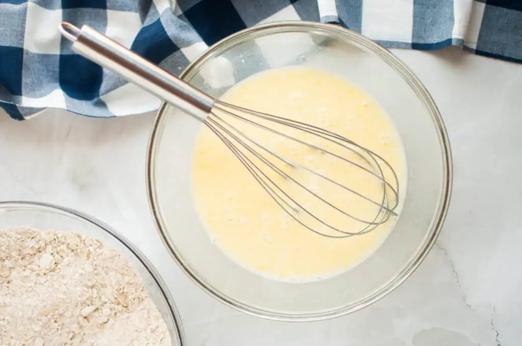 A mixing bowl with whisked eggs and milk next to a bowl of flour on a marble countertop with a blue and white checkered cloth nearby.