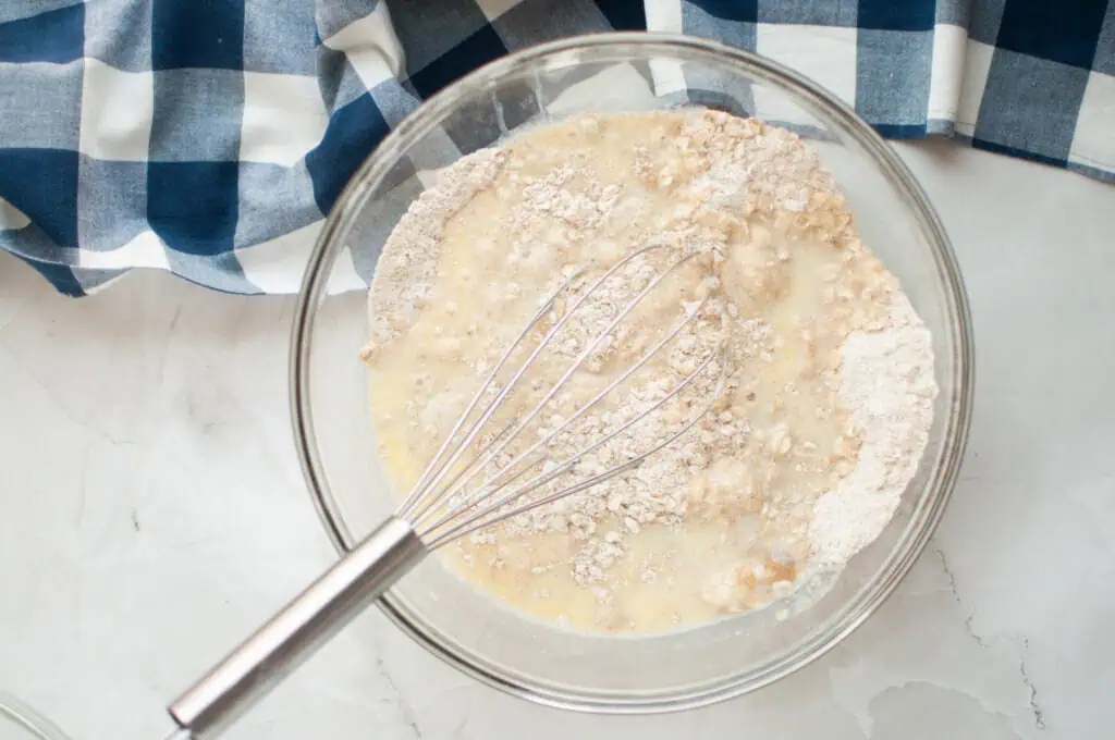A mixing bowl contains oat flakes, flour, and liquid being whisked together, with a blue and white checkered cloth nearby.