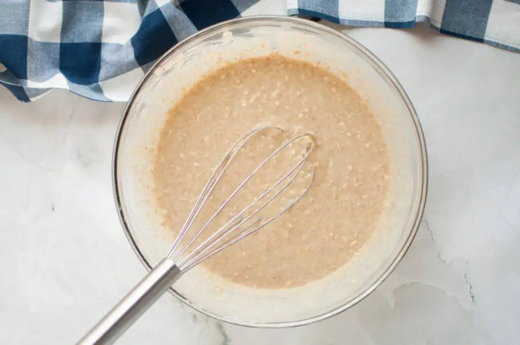 A whisk in a glass bowl with pancake batter, sitting on a light surface with a blue and white checkered cloth nearby.