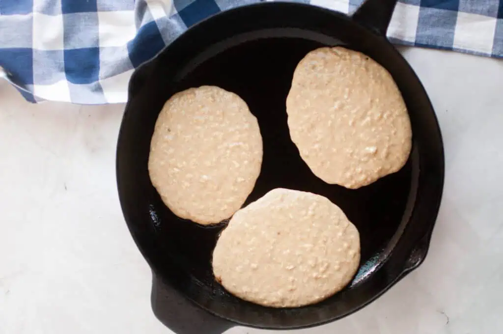 Three pancakes cooking in a black skillet on a marble surface, with a blue-and-white checkered cloth partially visible.