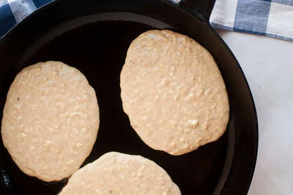 Three pancakes cooking in a black skillet, with a checkered cloth visible in the background.