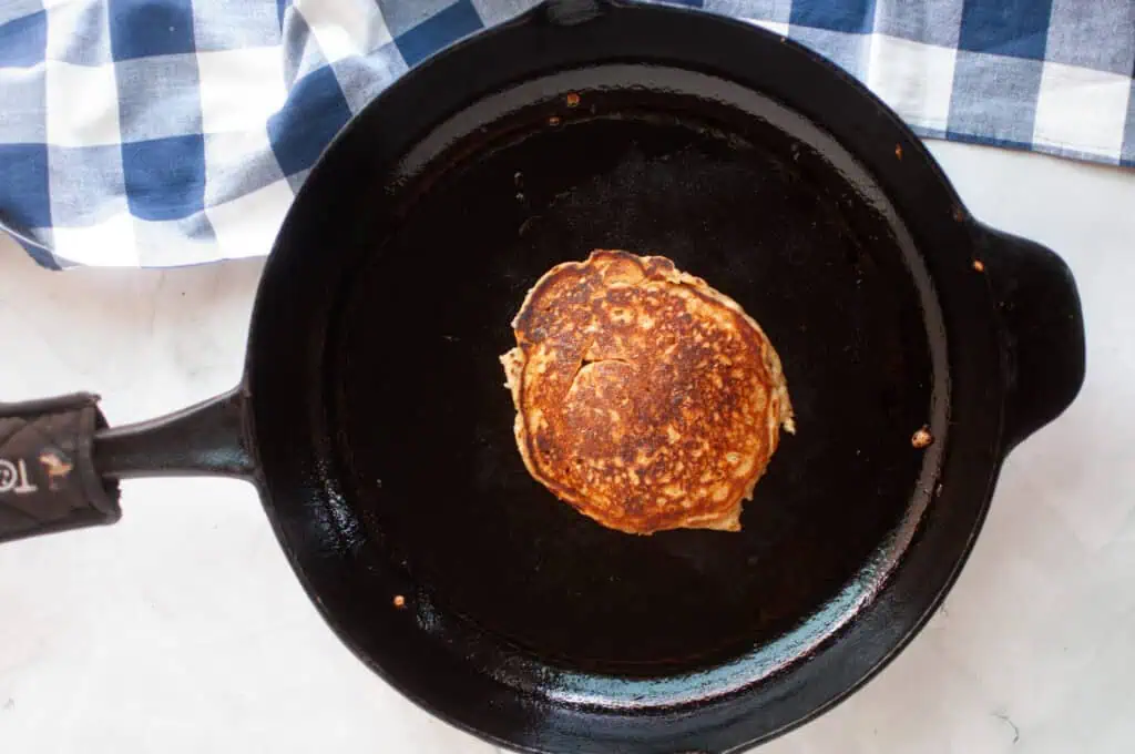 A pancake cooking in a black skillet, placed on a white surface with a blue and white checkered cloth nearby.