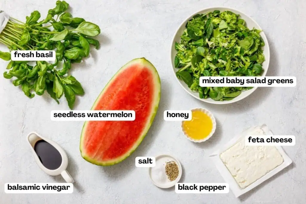 Ingredients for a salad arranged on a white surface: fresh basil, seedless watermelon, mixed baby salad greens, honey, feta cheese, balsamic vinegar, salt, and black pepper.
