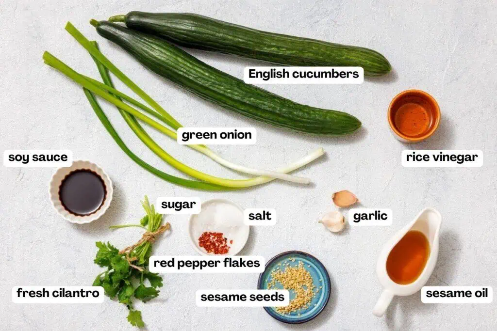Ingredients for a dish include English cucumbers, green onion, soy sauce, rice vinegar, sugar, garlic, sesame oil, sesame seeds, salt, red pepper flakes, and fresh cilantro on a table.
