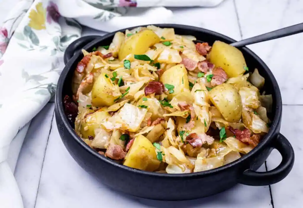 A black bowl filled with Cabbage & Potatoes with Bacon with a spoon on the side.