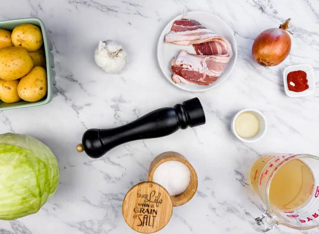 Ingredients for Cabbage & Potatoes with Bacon on a marble surface include potatoes, cabbage, garlic, bacon, onion, salt, pepper, smoked paprika, olive oil, and a measuring cup with liquid.