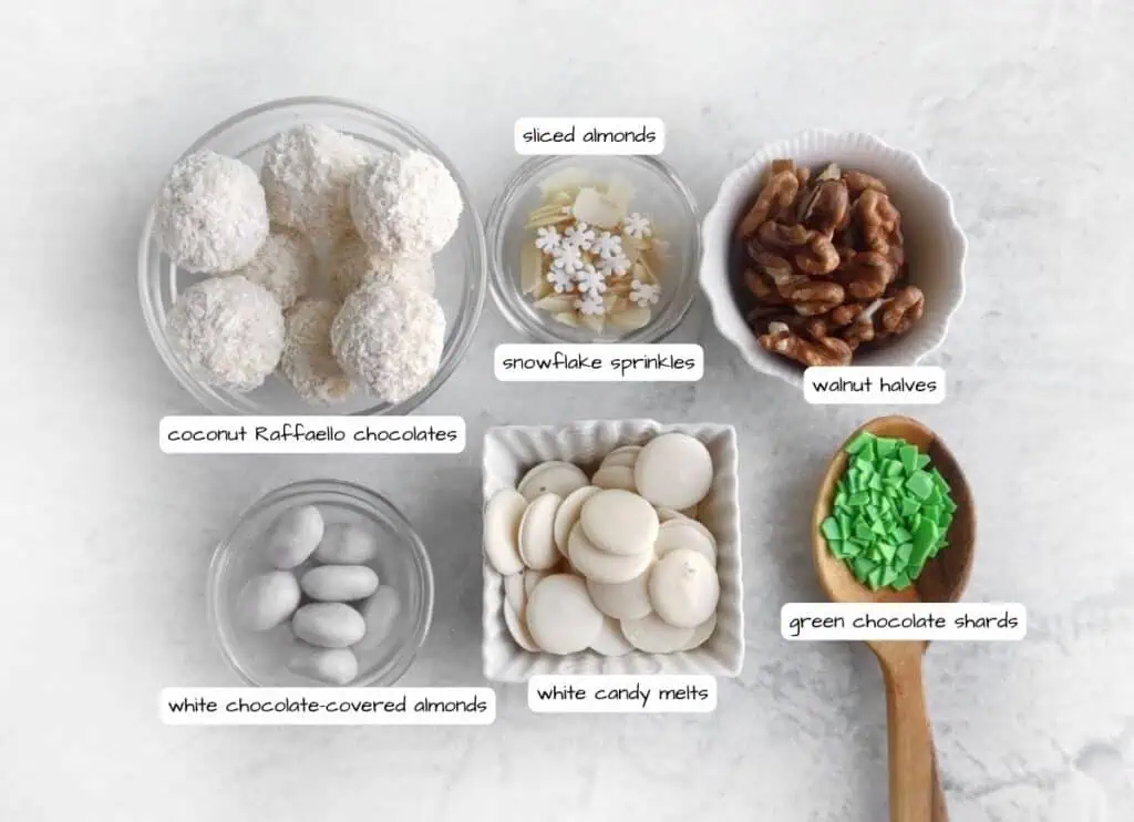 Various sweet toppings and ingredients displayed in bowls: coconut chocolates, white chocolate-covered almonds, sliced almonds, walnut halves, snowflake sprinkles, white candy melts, and green chocolate shards.