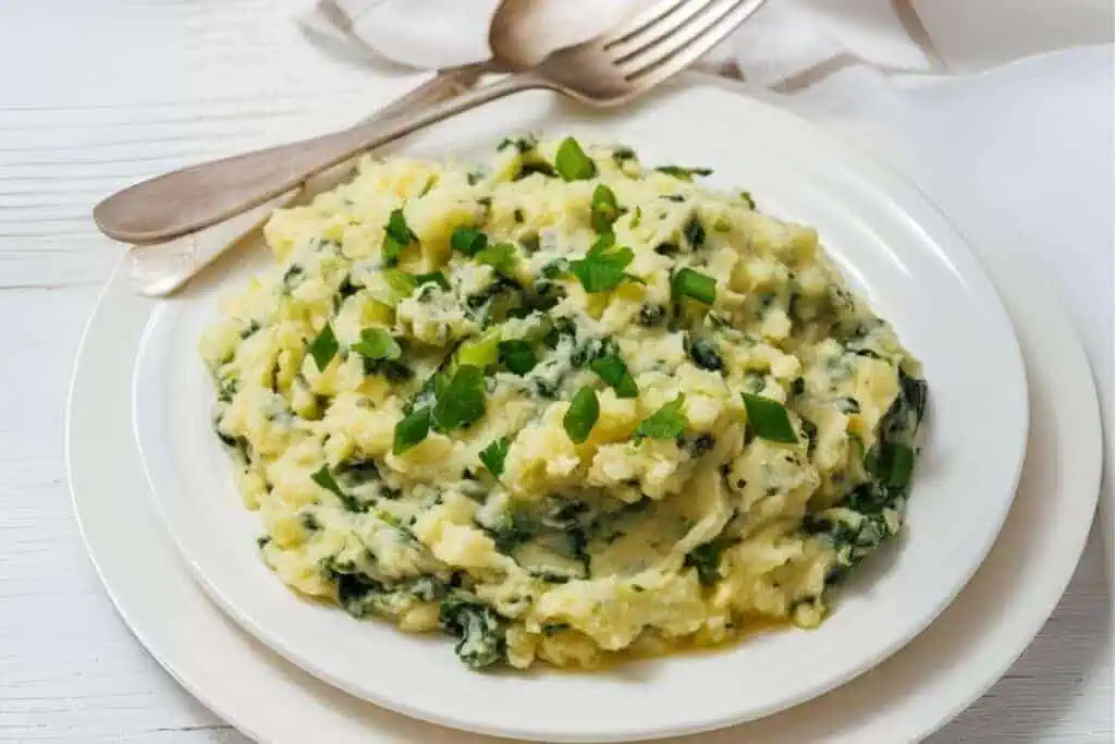A white plate with colcannon and kale with green herbs is garnished with chopped scallions