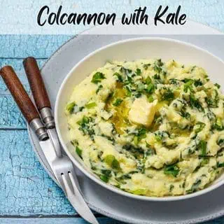 A bowl of Colcannon with Kale with parsley and melting butter on top, placed on a gray plate with a fork and knife on a rustic blue wooden table.
