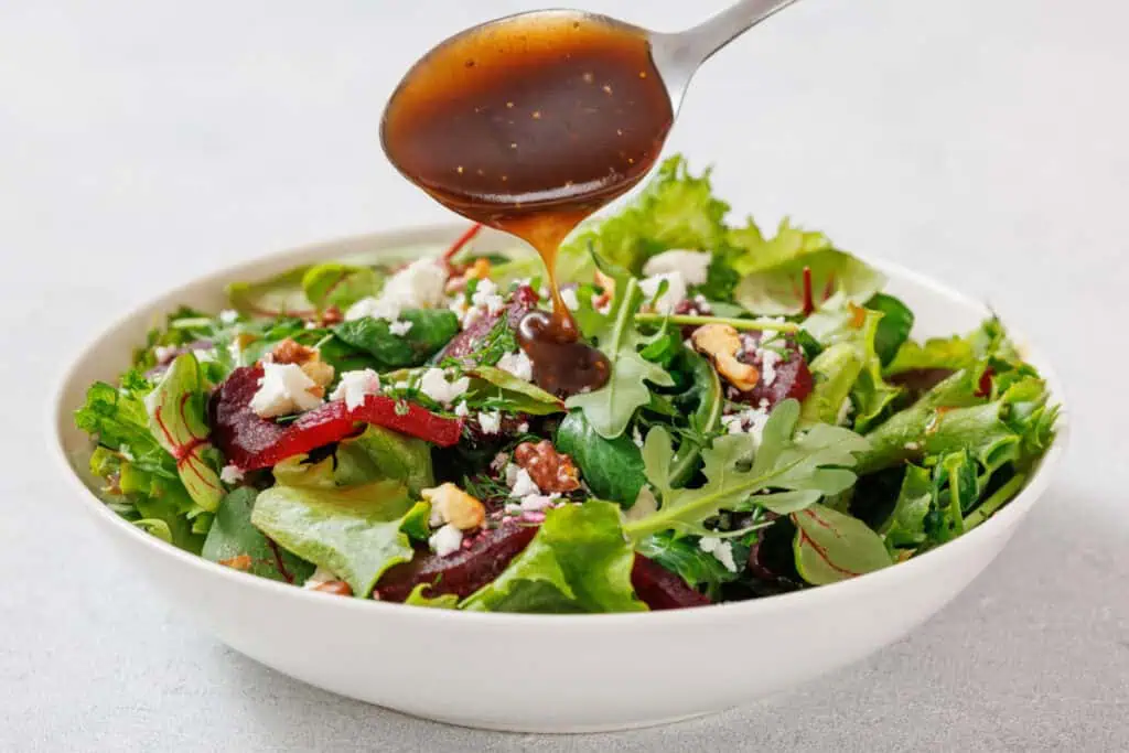 A spoon drizzles dark dressing over a salad in a white bowl containing mixed greens, beet slices, feta cheese, and walnuts.