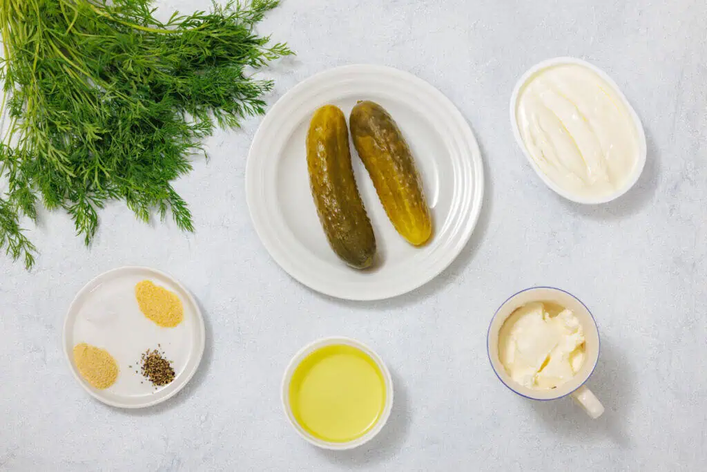 Pickles on a white plate surrounded by fresh dill, sour cream, mayonnaise, oil, salt, pepper, and garlic powder on separate dishes.