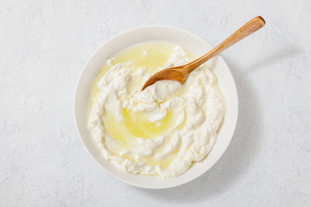 A bowl of yogurt with olive oil, containing a wooden spoon, placed on a light textured surface.