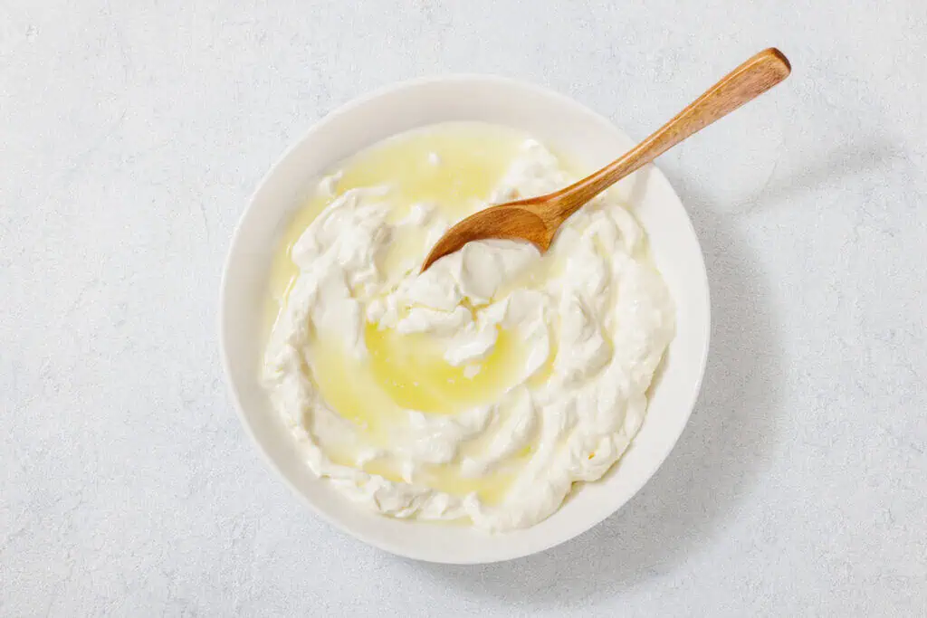 A bowl of yogurt with olive oil, containing a wooden spoon, placed on a light textured surface.