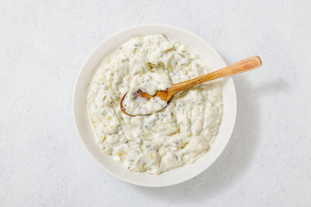 A bowl of creamy tzatziki sauce with herbs, shown from above. A wooden spoon rests in the sauce.