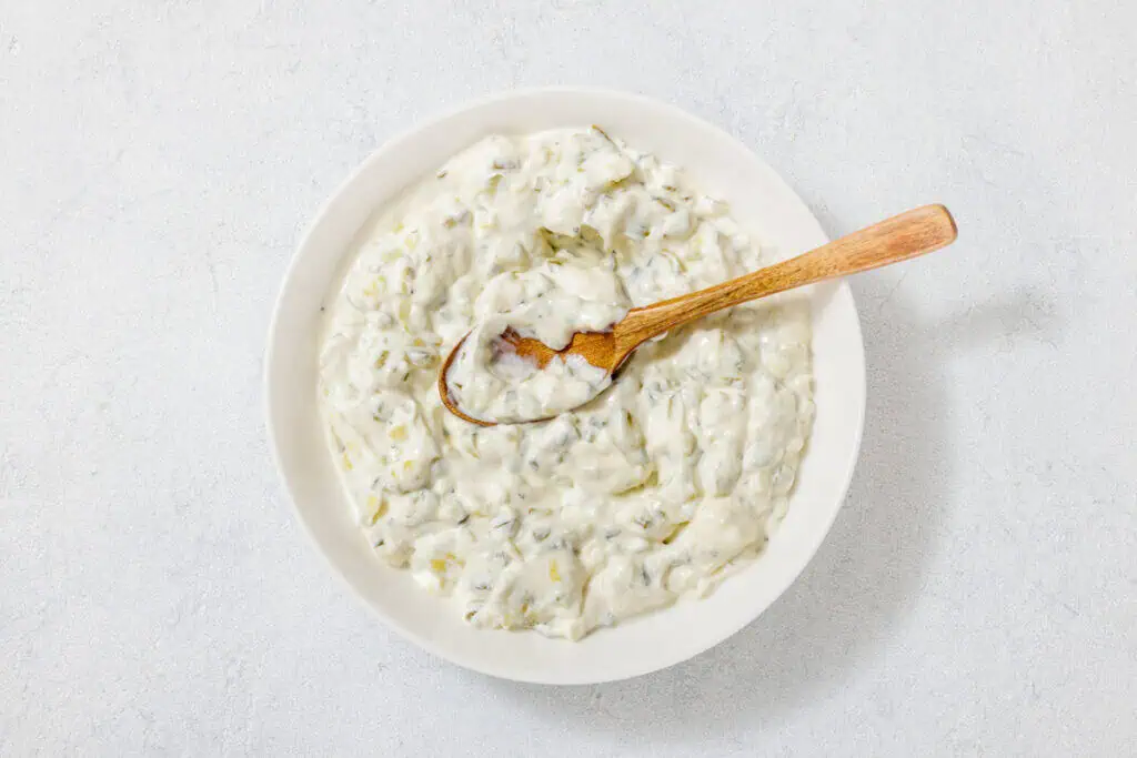 A bowl of creamy tzatziki sauce with herbs, shown from above. A wooden spoon rests in the sauce.