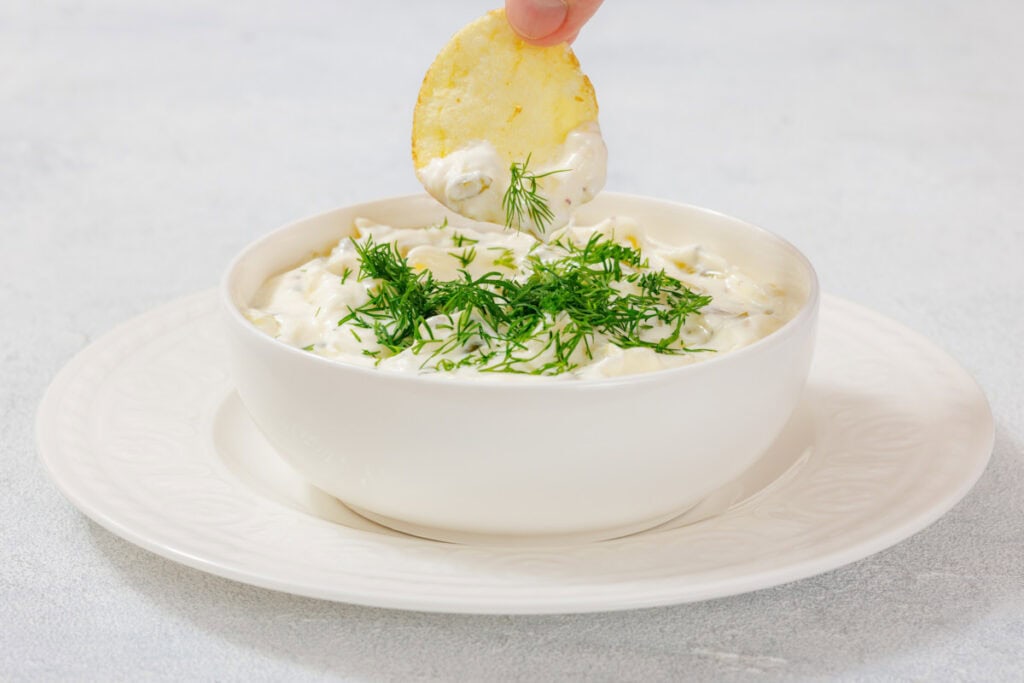 A hand dips a potato chip into a white bowl of creamy Dill Pickle Dip garnished with fresh dill sprigs.