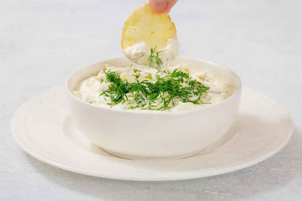 A hand dips a potato chip into a white bowl of creamy Dill Pickle Dip garnished with fresh dill sprigs.
