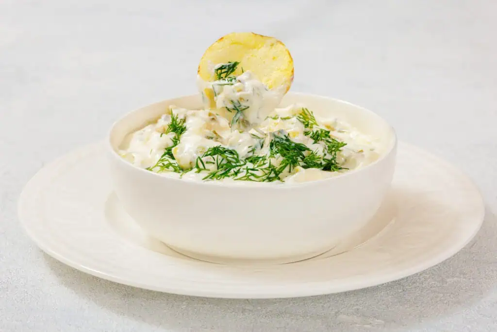 A bowl of creamy Dill Pickle Dip topped with chopped dill, with a potato chip dipped into it, placed on a white plate.