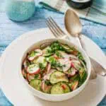 Bowl of Radish Salad with herbs on a white plate. Vintage fork and spoon on the side. Light blue wooden table background with a small jug and a napkin.